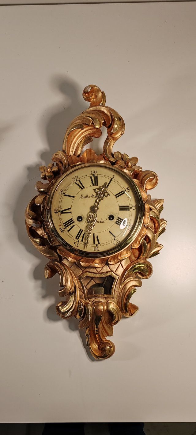 Emil N. Stockholm -seinäkello, jossa on lehtikultaus koristeissa. Korkeus 53cm ja leveys 29cm